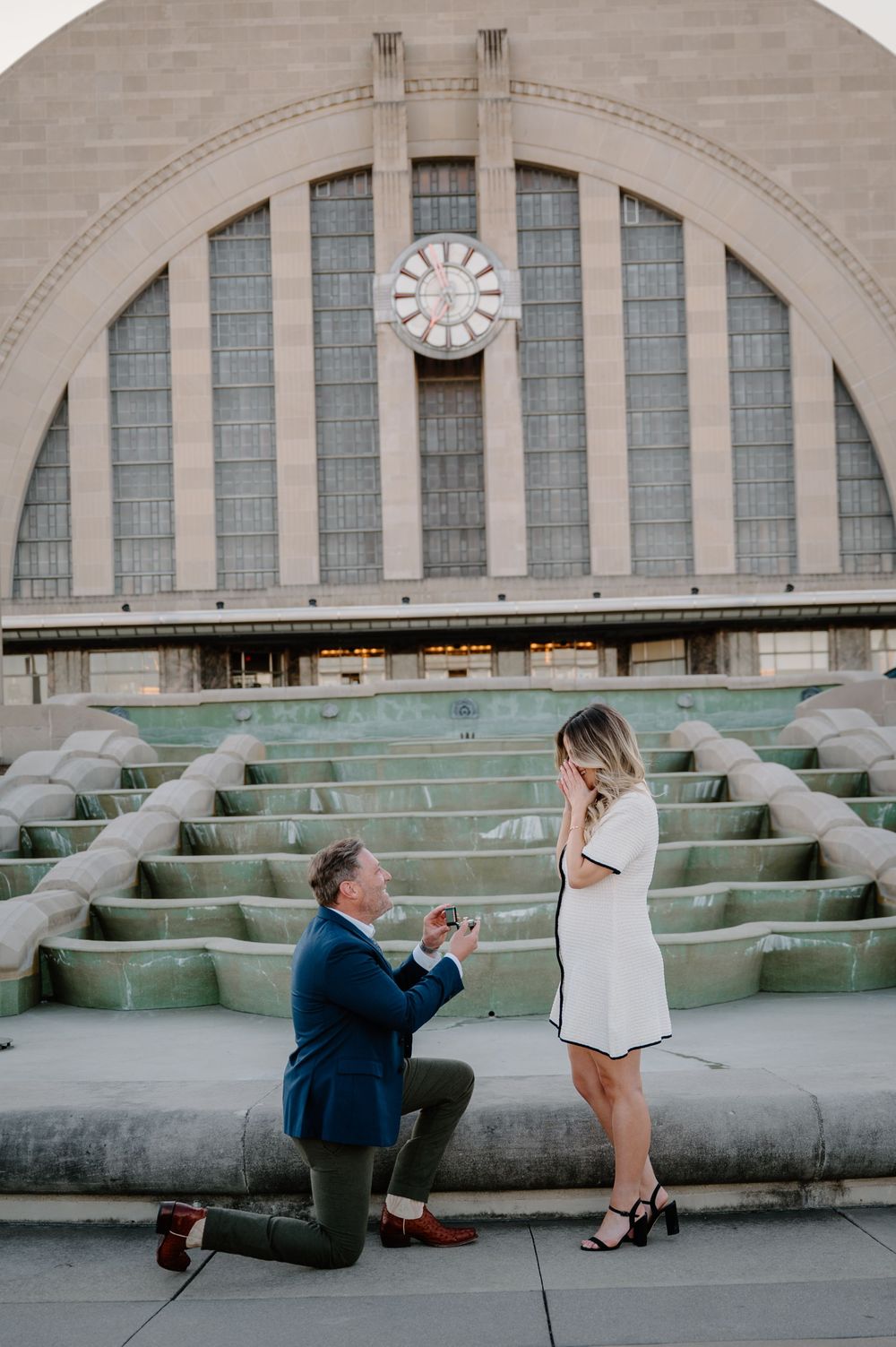 Man proposes on one knee to woman in white dress at Cincinnati Union Terminal fountain.