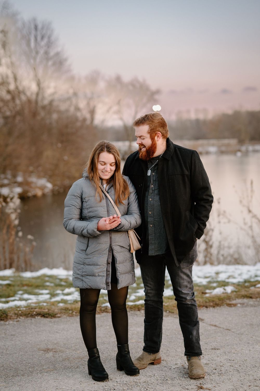 A smiling couple in winter coats stands near a snowy lakeside at sunset, the woman looking at her phone.