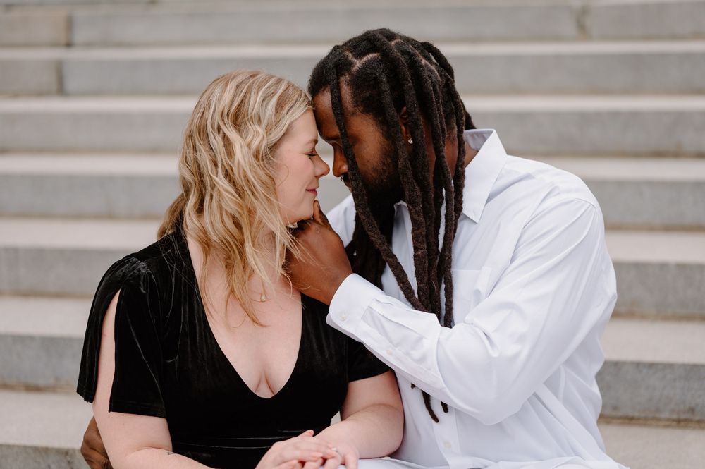 Romantic interracial couple sharing an intimate moment on stone steps, woman in black velvet dress, man in white shirt with dreadlocks.