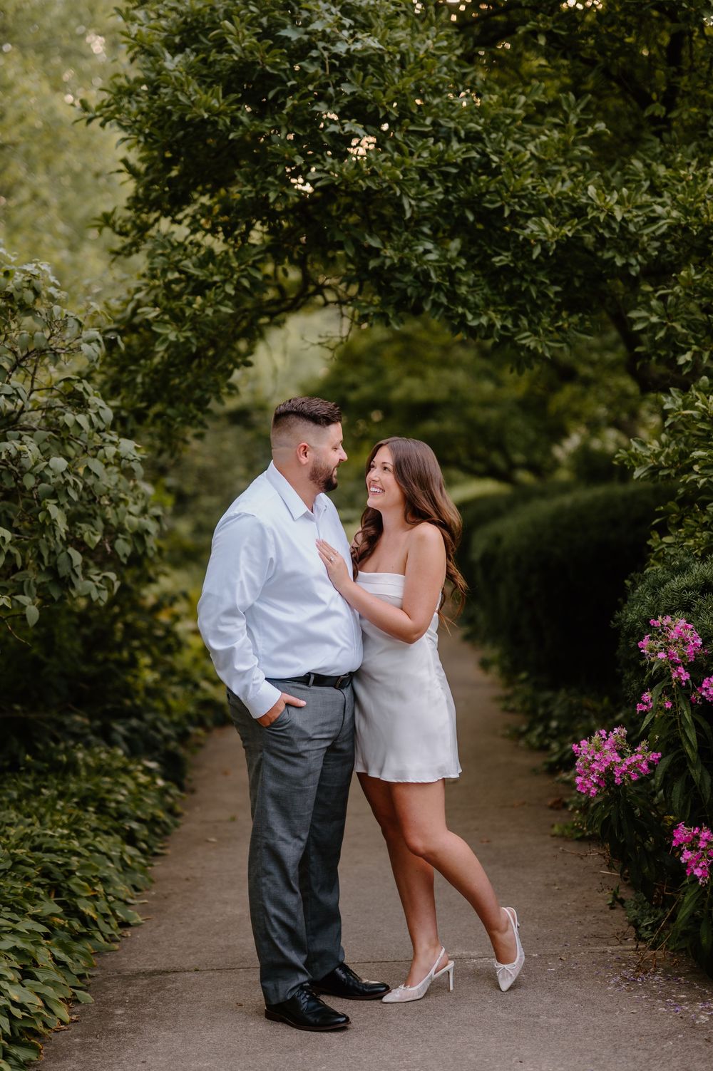 A romantic couple embraces in a serene garden path lined with lush greenery and blooming pink flowers.