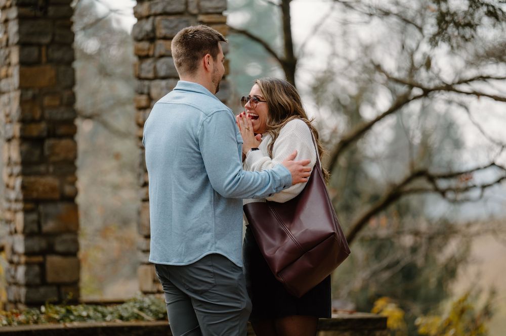 Couple sharing a romantic moment outdoors near stone pillars, woman with glasses holding a burgundy bag looks surprised.