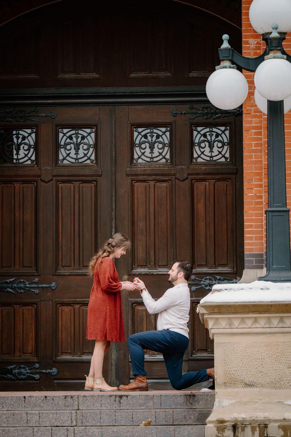 Man kneeling on one knee proposing to woman in red dress on ornate building steps with wooden doors.