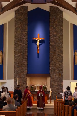 Catholic Mass being celebrated in a modern church with stone columns and a large illuminated crucifix on blue background.
