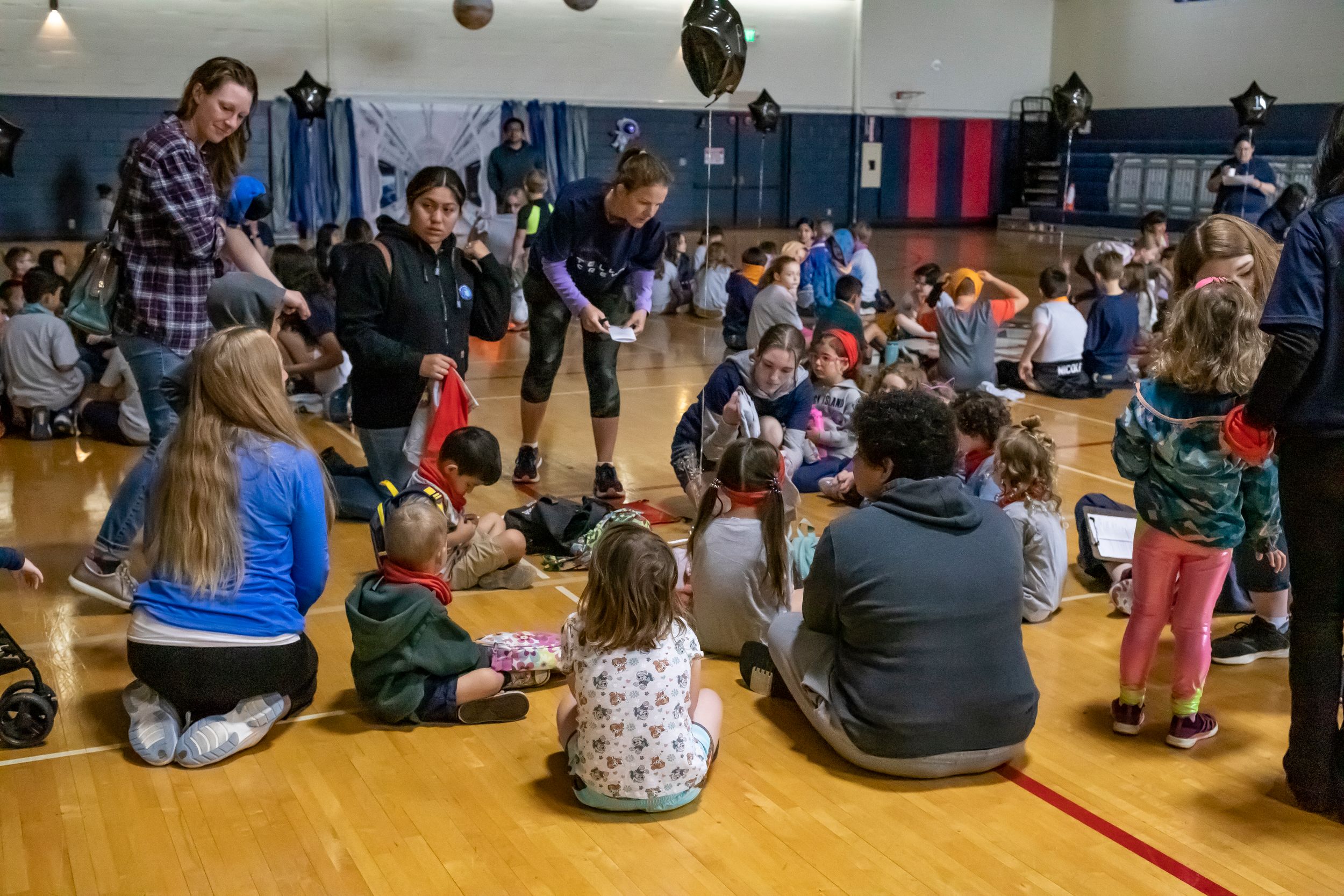 Children and adults sitting in circles during an interactive workshop or activity in a school gymnasium.