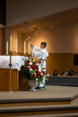 An altar server lights candles during a church service surrounded by red and white floral arrangements.
