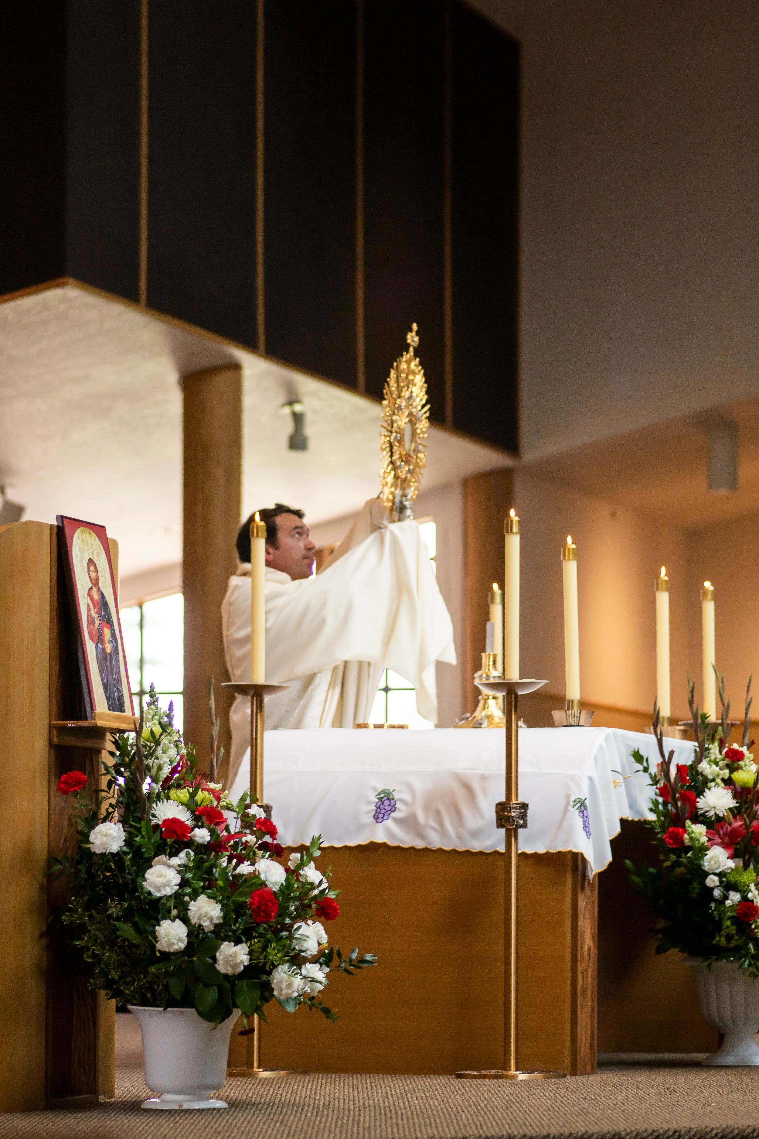 A priest raises a golden monstrance during a Catholic mass service with floral decorations on the altar.