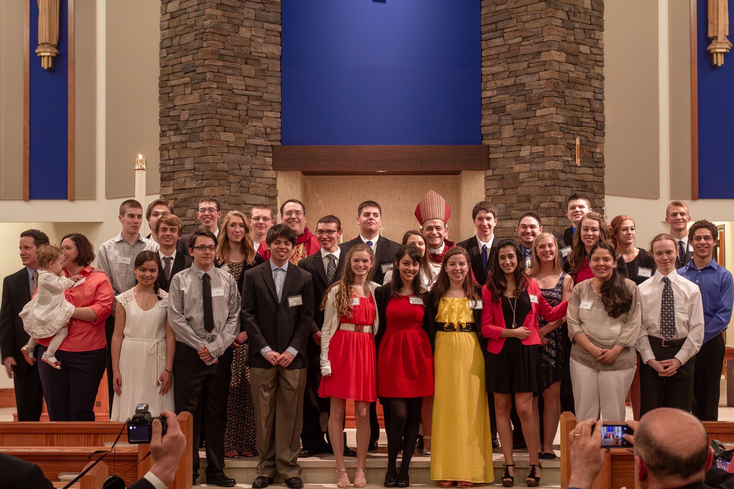 A formal group photo of young people in dress attire standing together in front of a stone church wall and blue backdrop.
