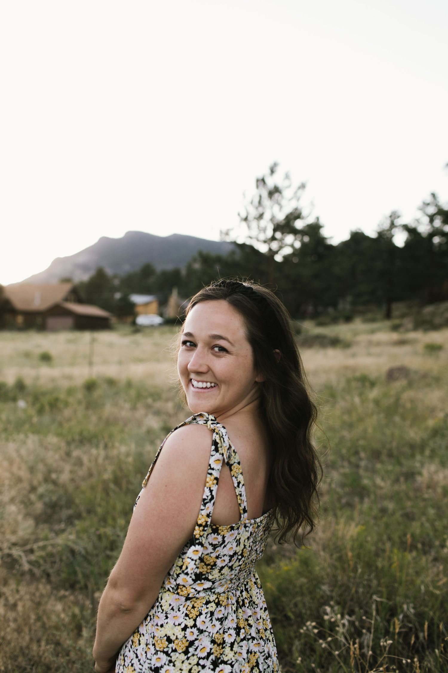 A woman in a floral dress smiles over her shoulder in a field at sunset with mountains in background.