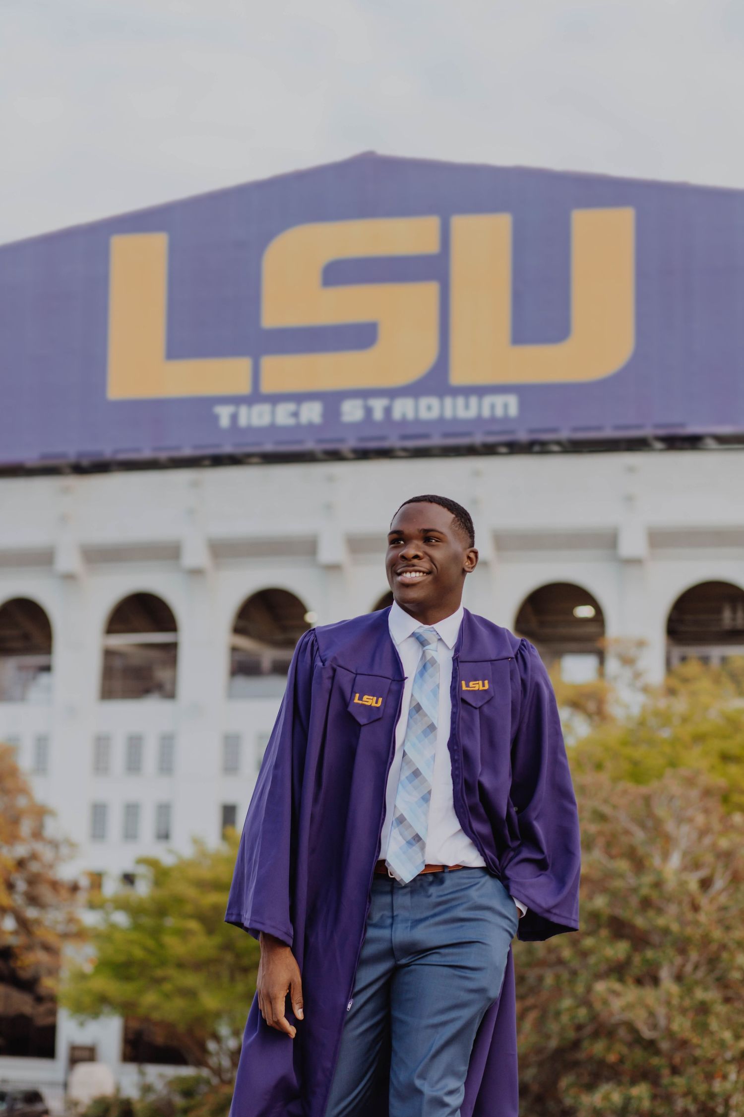A graduate in purple academic regalia poses in front of LSU Tiger Stadium.