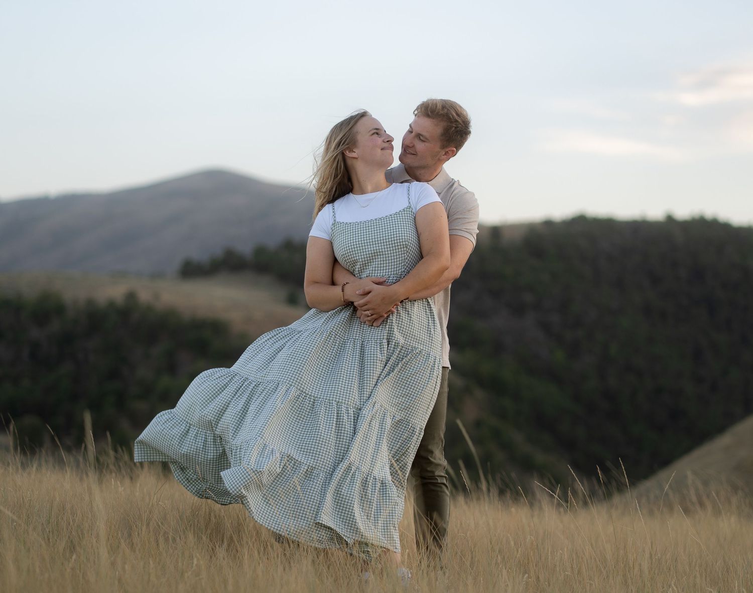 Engaged couple holding each other while the wind blows in Cache Valley. Looking into each others eyes with sprawling hills in the back.