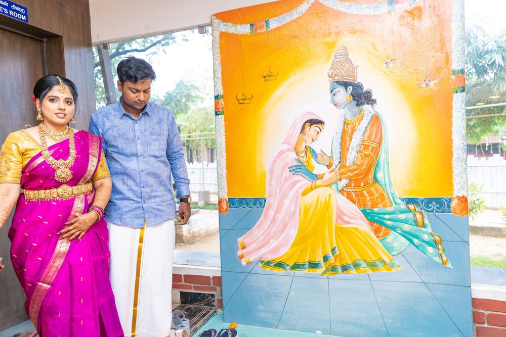 A couple poses next to a colorful painting of Krishna and Radha against an orange backdrop.
