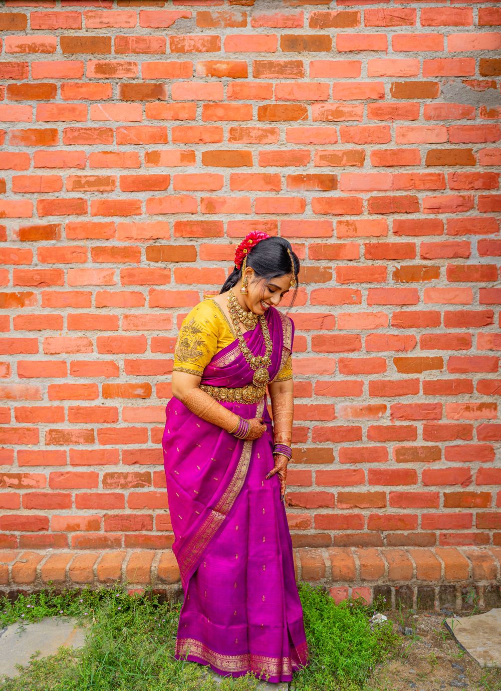 Woman in bright pink and yellow sari poses against red brick wall with grass below.