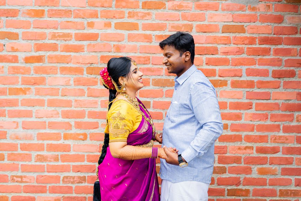 A couple in traditional Indian attire pose lovingly against a brick wall, with her in pink and yellow sari and him in blue shirt.