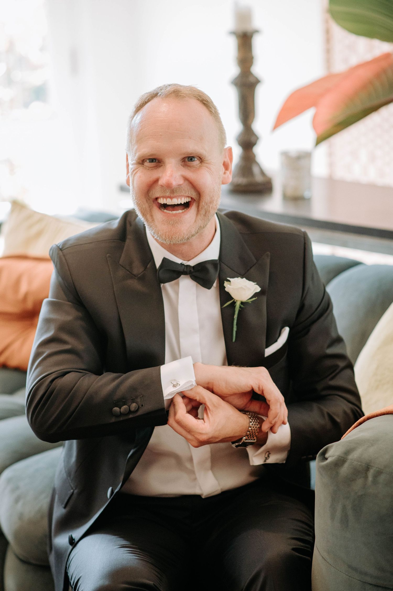 A formal portrait of someone in a classic black tuxedo sitting on a grey couch.