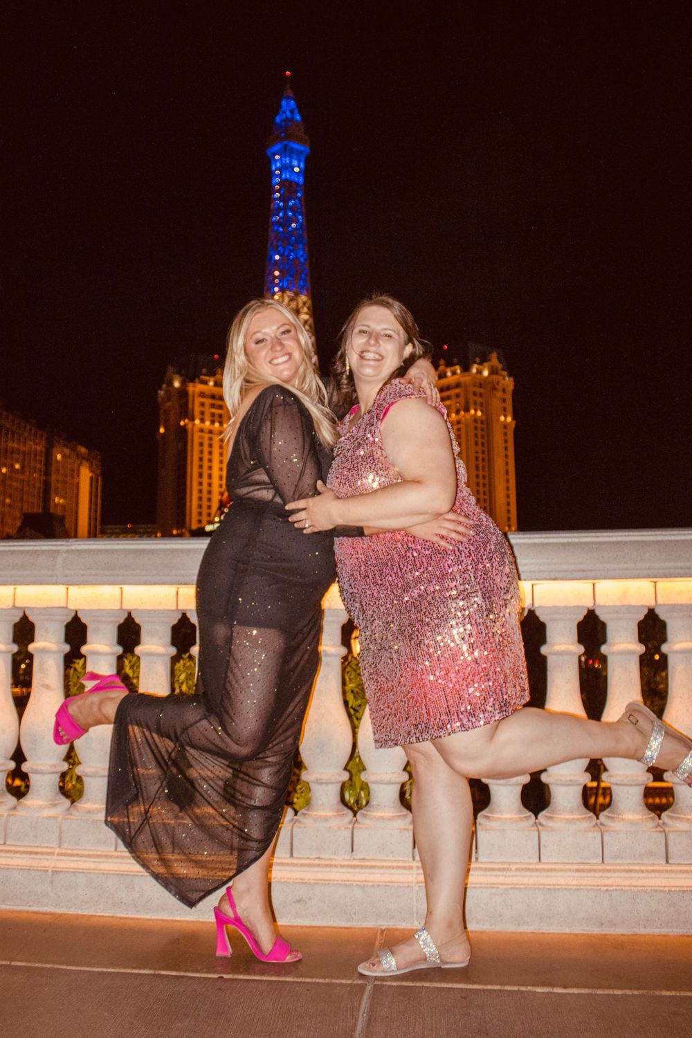 Two women posing and smiling in front of the Paris Las Vegas Eiffel Tower replica at night.