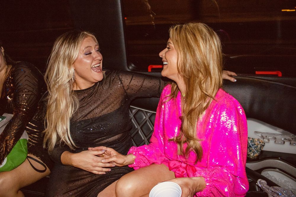 Two women laughing together in a limo, one in a black dress and one in a pink sequin outfit.