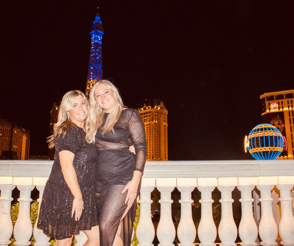 Two women posing in front of the Paris Las Vegas Eiffel Tower at night.