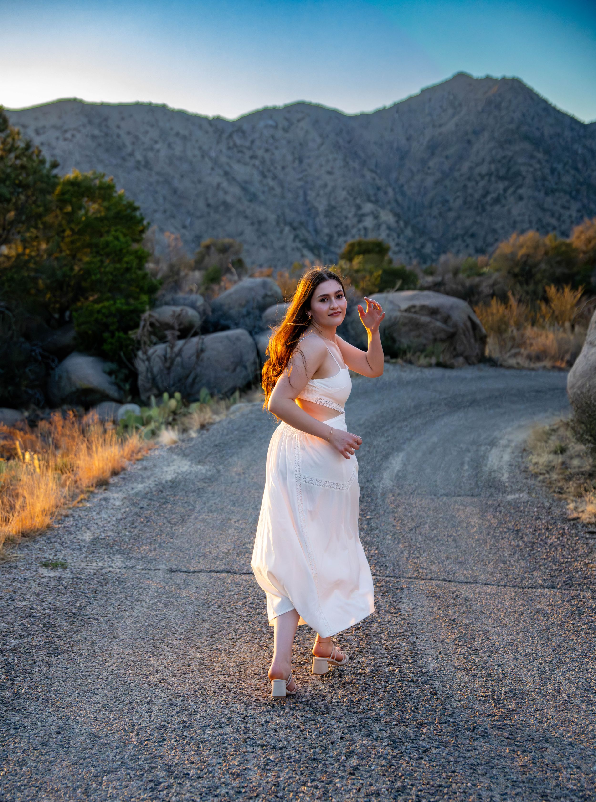 A graceful figure in a white dress poses on a mountain dirt road at golden hour with mountains in the background.