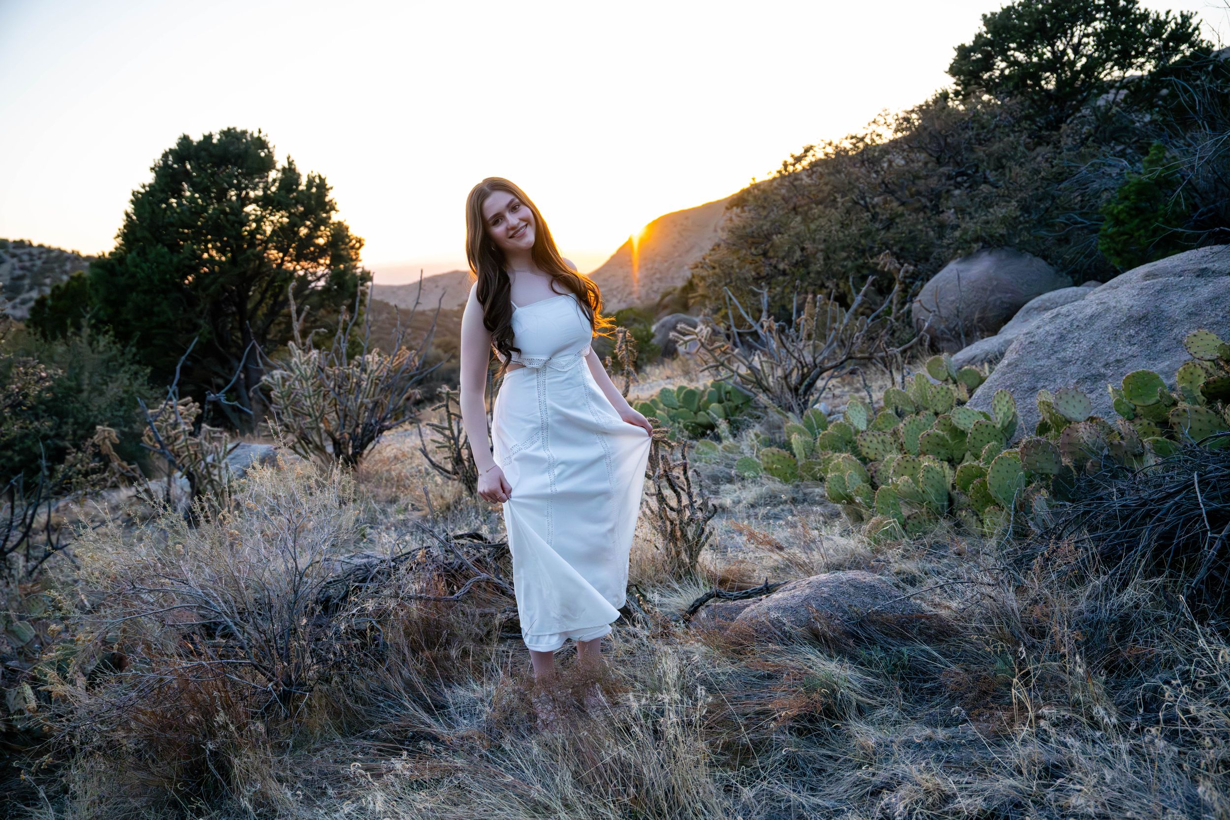 A person in a flowing white dress poses among desert rocks during golden hour sunset.