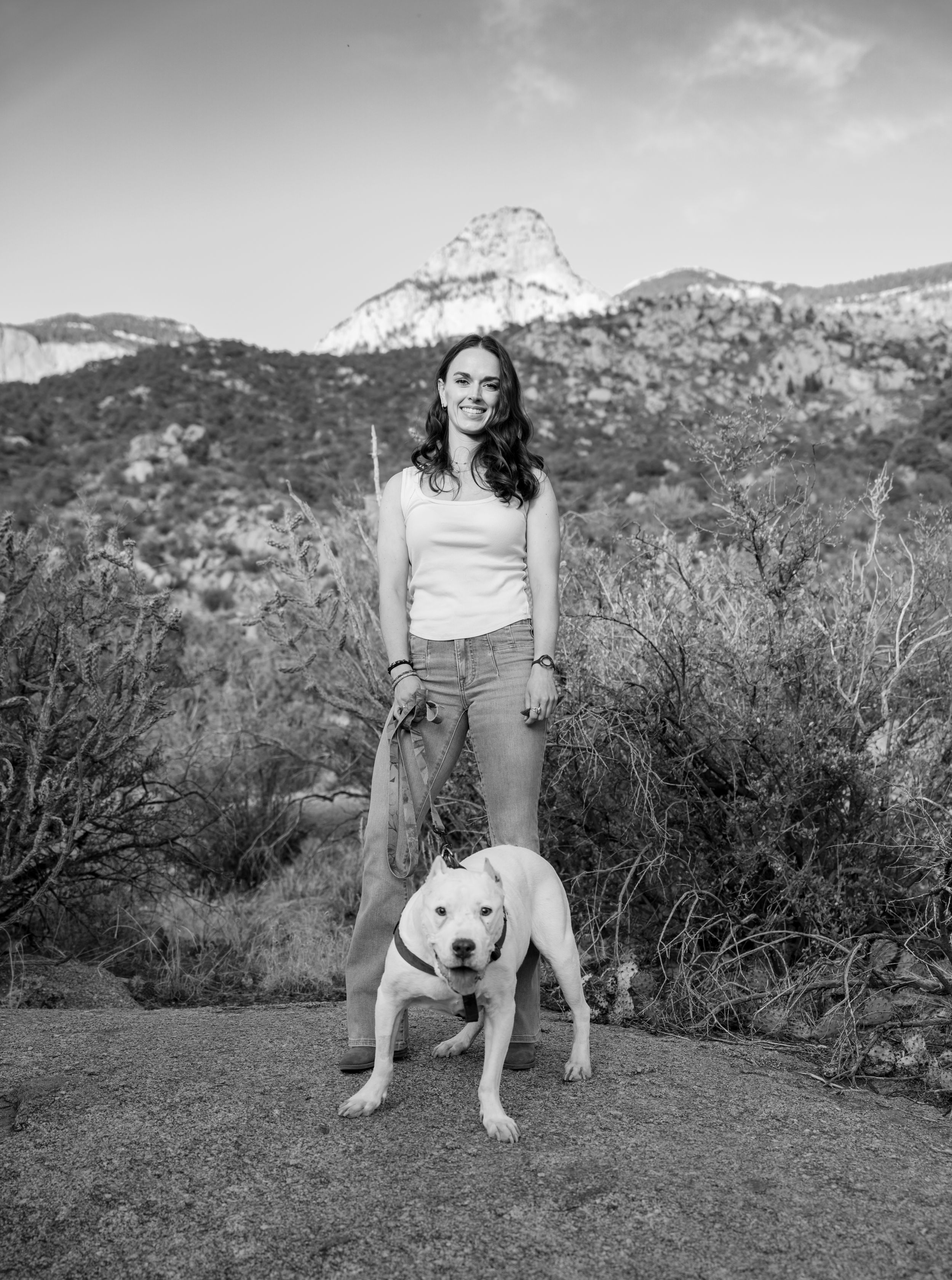 A black and white photograph shows someone standing outdoors with a white dog against a mountainous desert landscape.