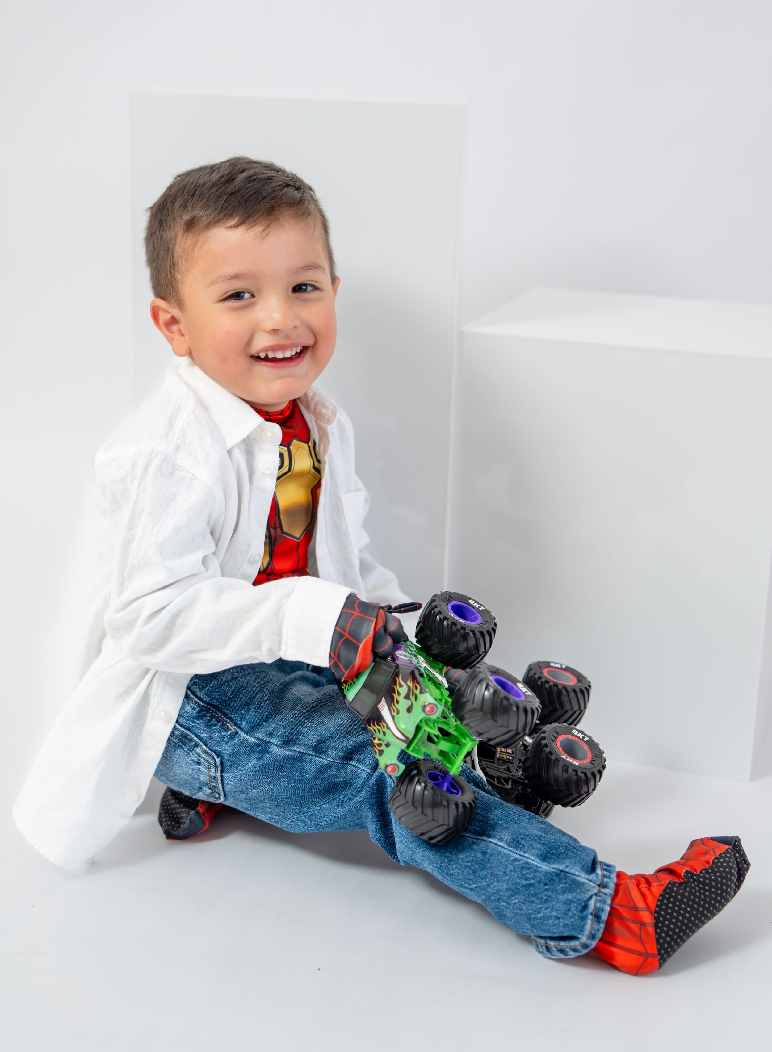 A joyful child wearing a white shirt and tie sits on the ground with colorful toy monster trucks.