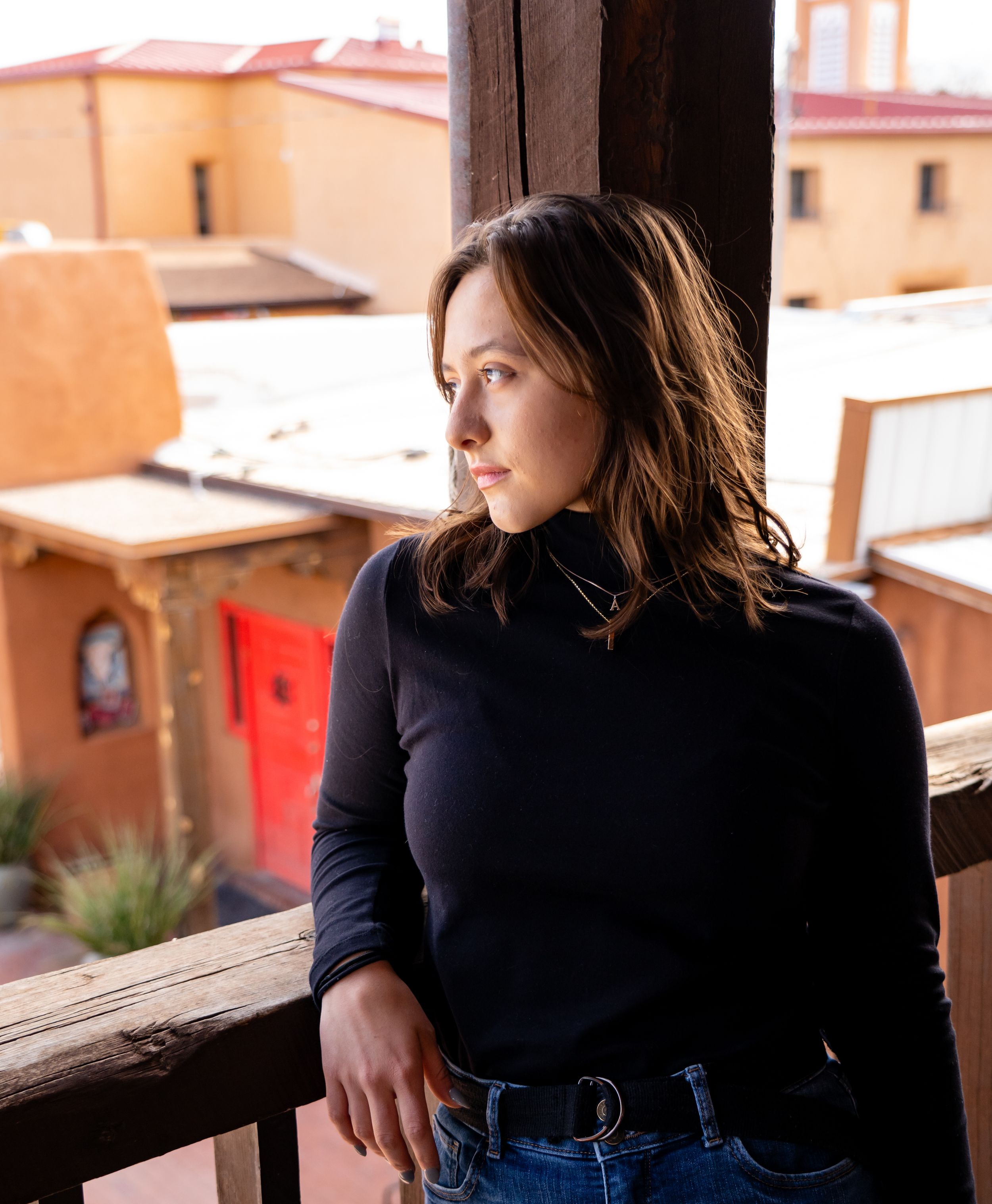 Woman in black sweater gazes thoughtfully from balcony overlooking adobe buildings.