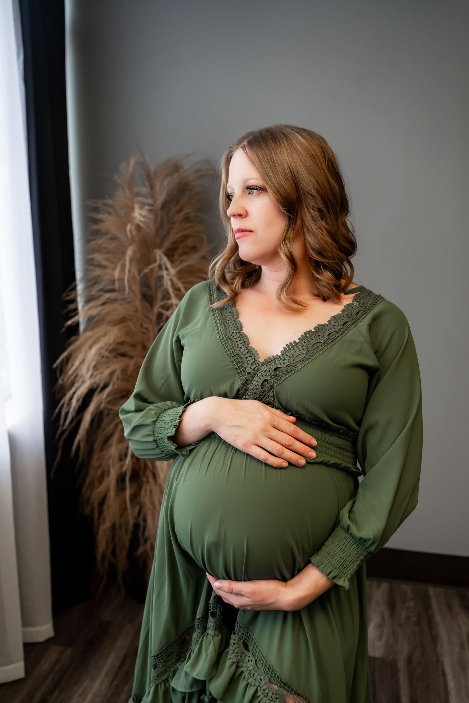 Intimate maternity portrait in olive green lace dress by window with pampas grass.