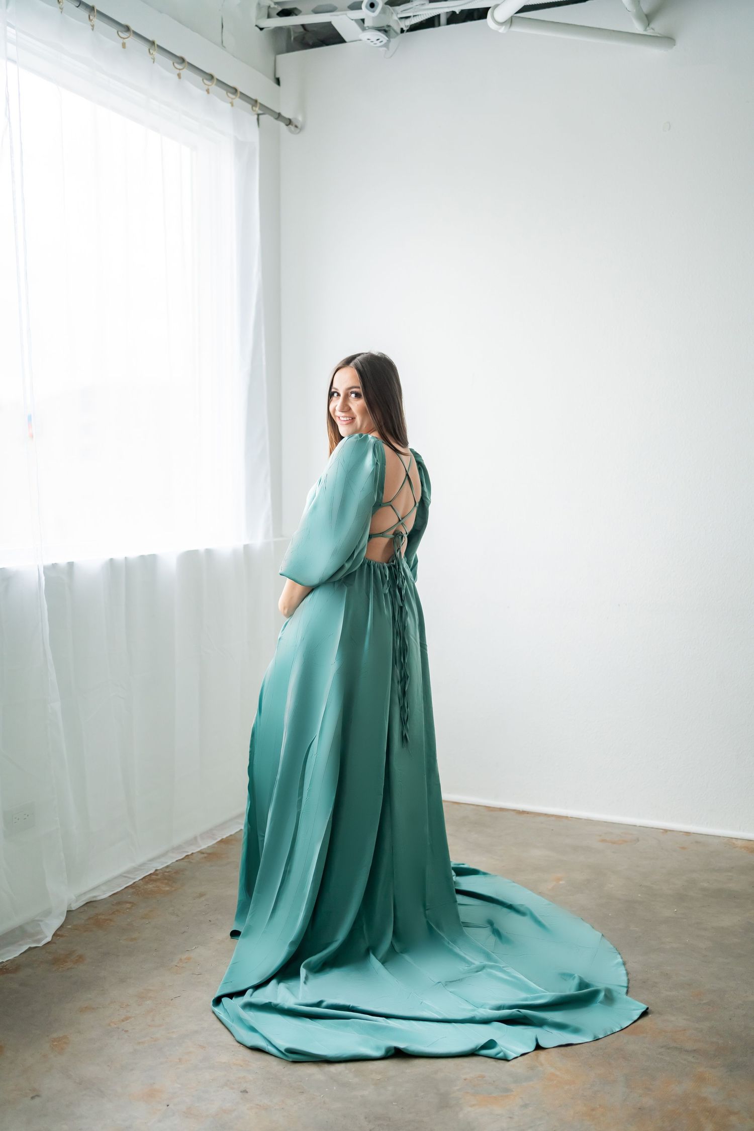Back view of a teal maternity gown with dramatic train and lace-up detail in a minimalist setting.