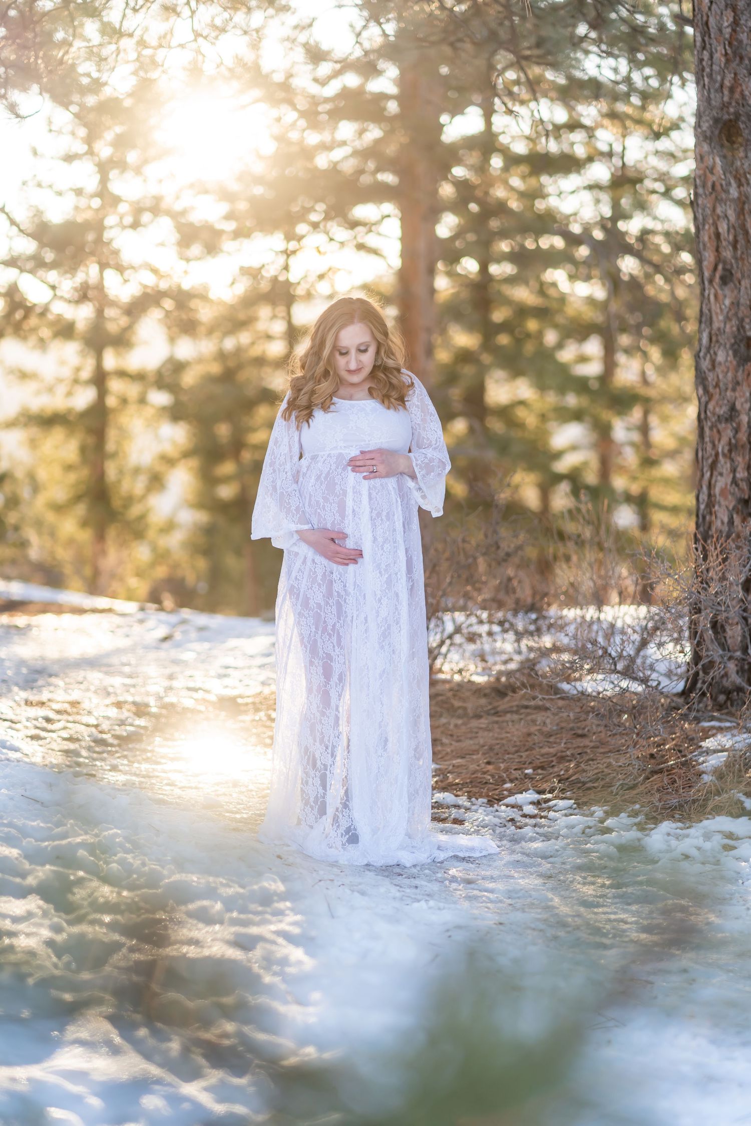 A winter maternity photo with a white lace dress against snowy forest backdrop at sunset.