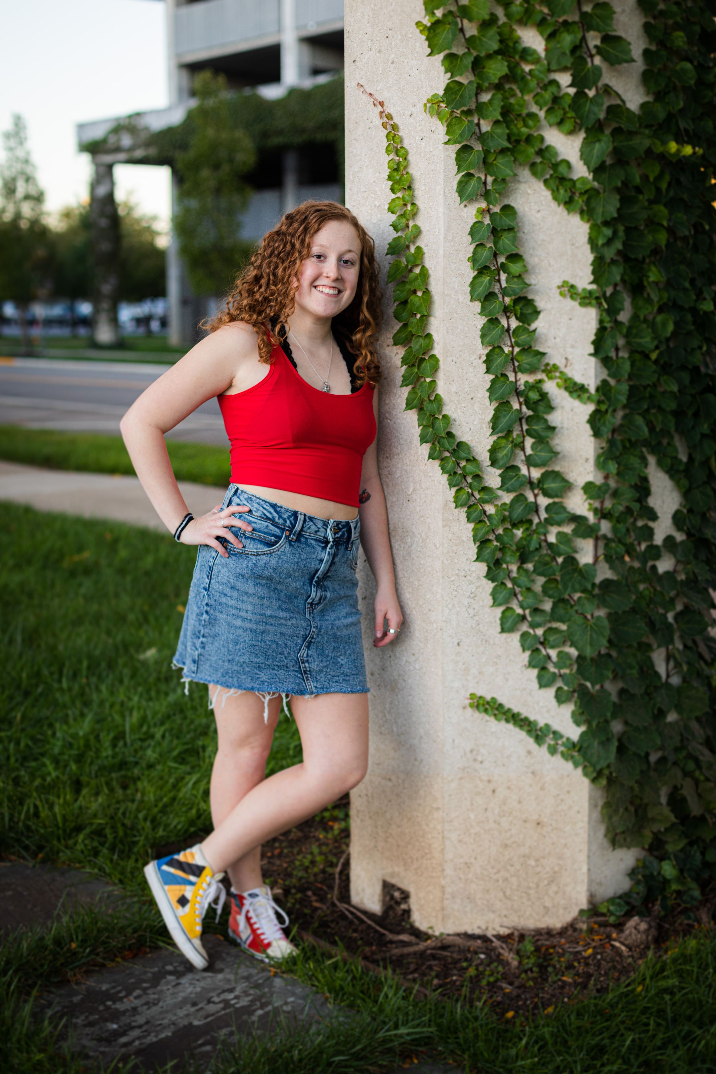 Person in red top and denim skirt poses near ivy-covered wall outdoors.