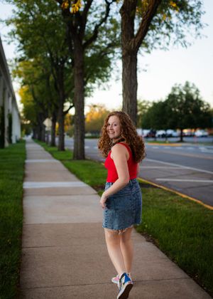 Person with curly hair in casual attire poses energetically on a suburban sidewalk.