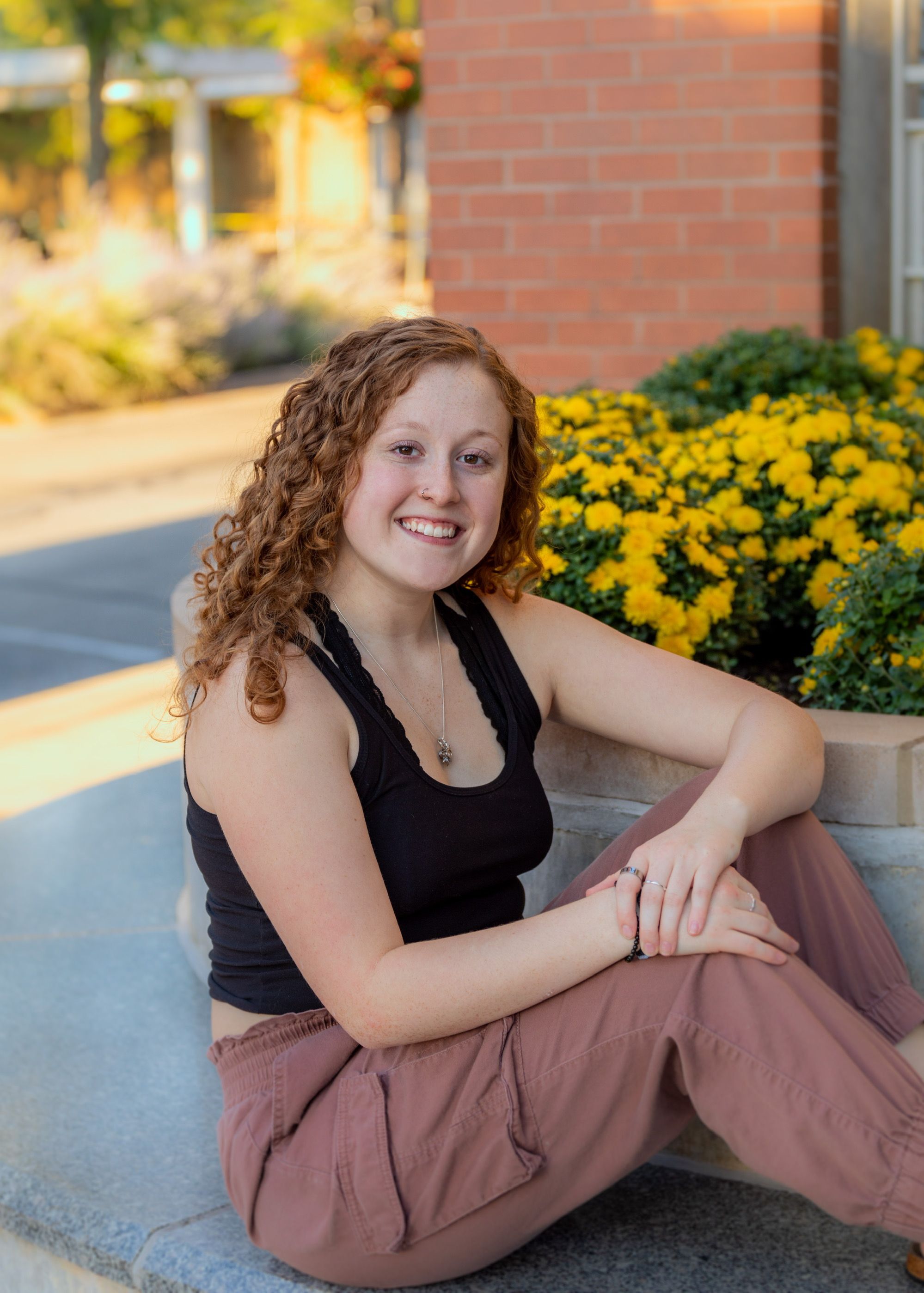Casual portrait by brick wall and yellow flowers wearing black top and brown pants.