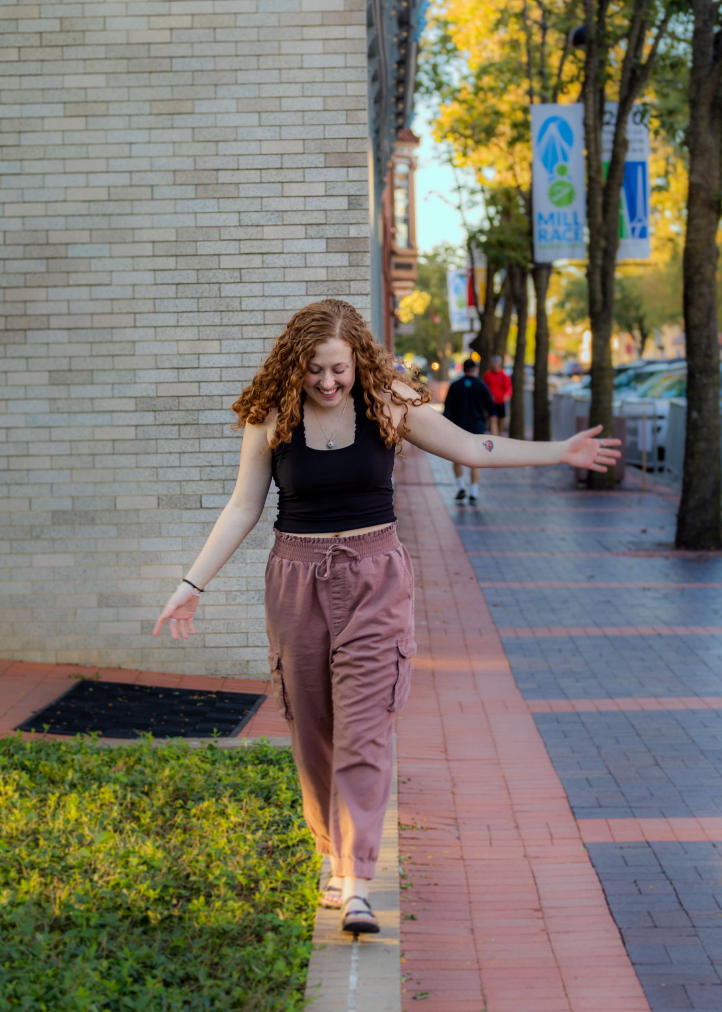 Person in black top and mauve pants poses playfully on a brick-lined sidewalk with trees.