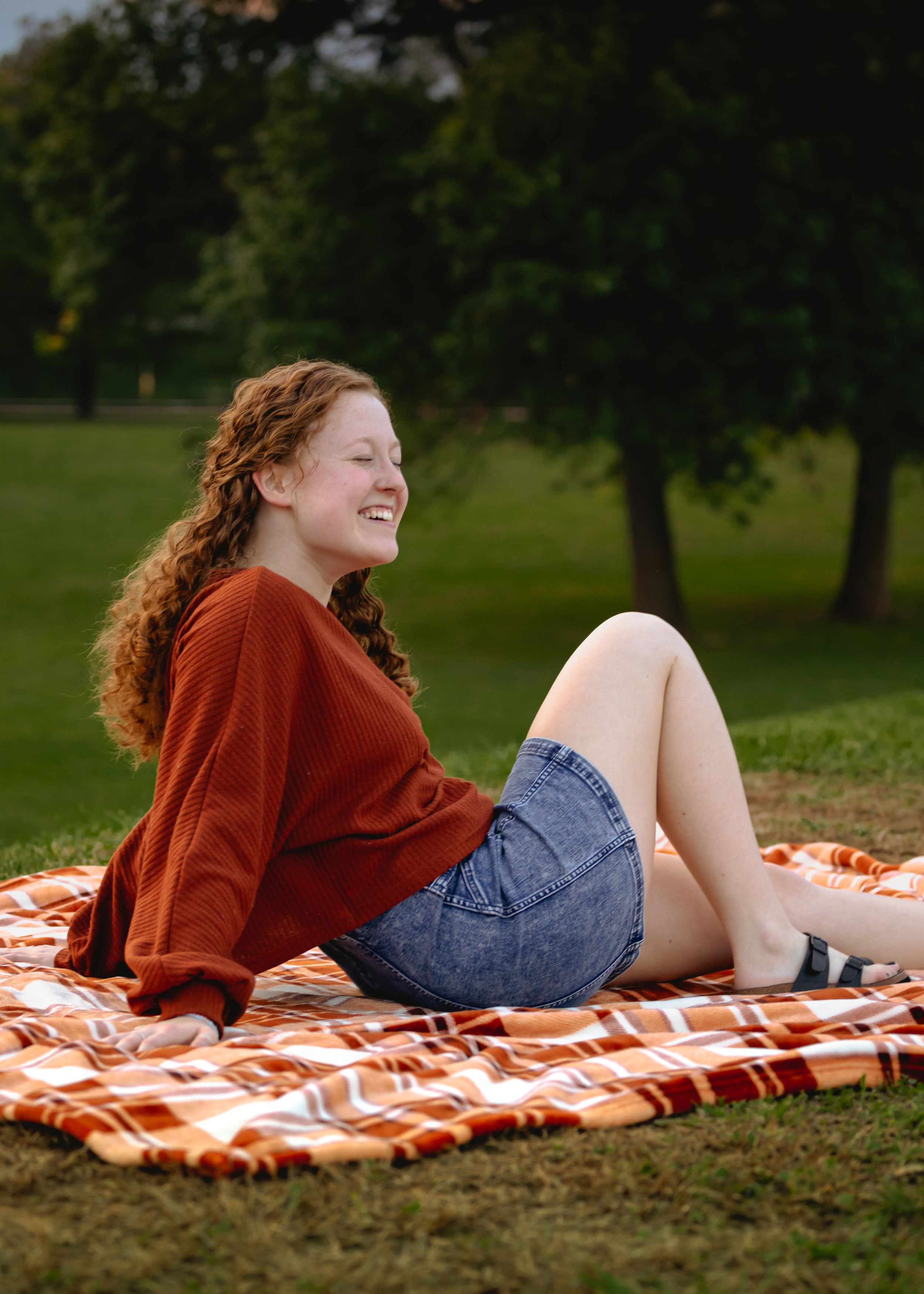 Person relaxing on orange striped blanket in park wearing burgundy sweater and jeans.