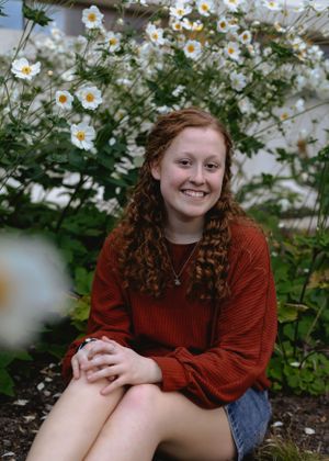 Someone posing near white daisy flowers wearing a red sweater and denim shorts.