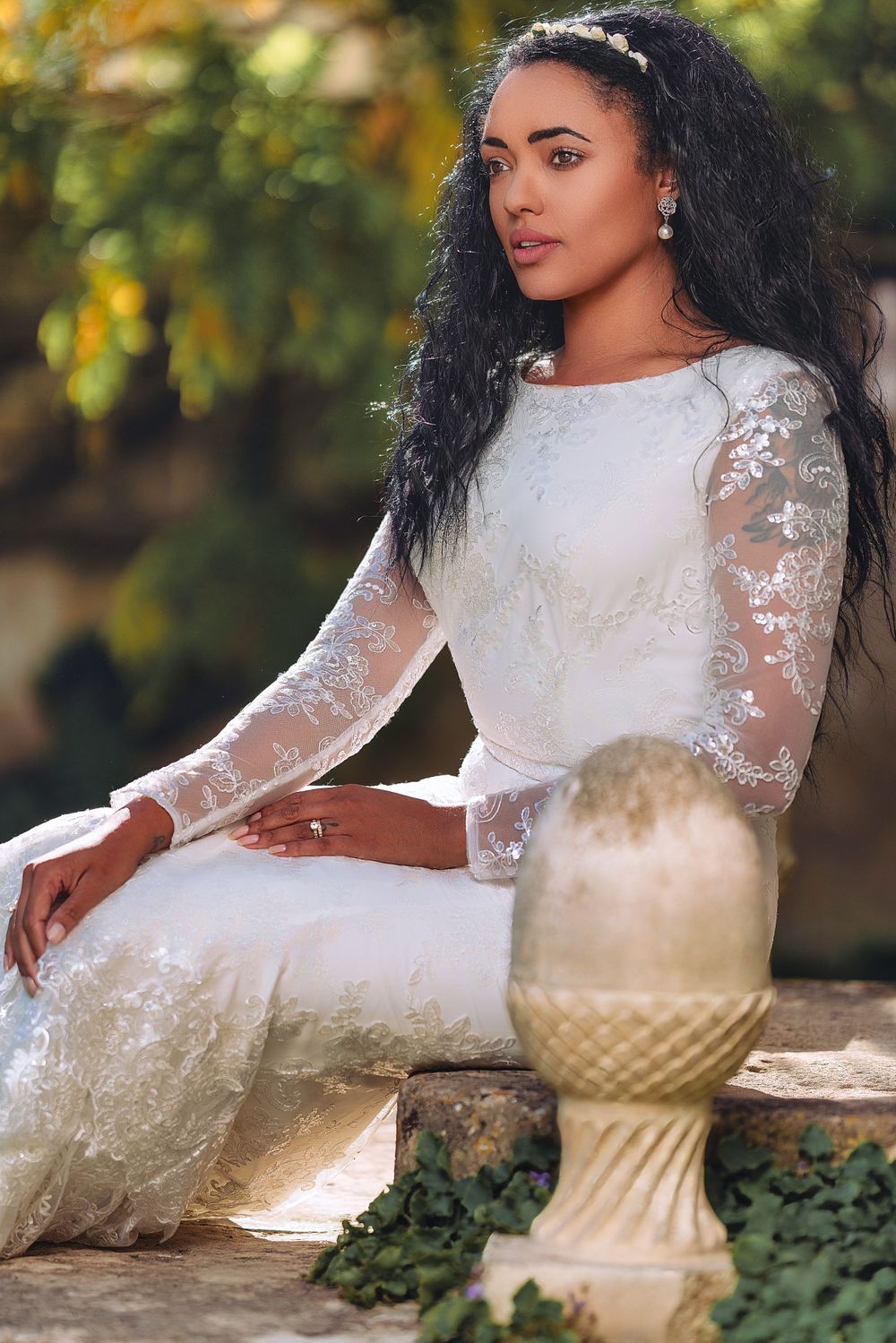 Woman in white lace dress poses elegantly against outdoor garden setting with decorative pedestal.