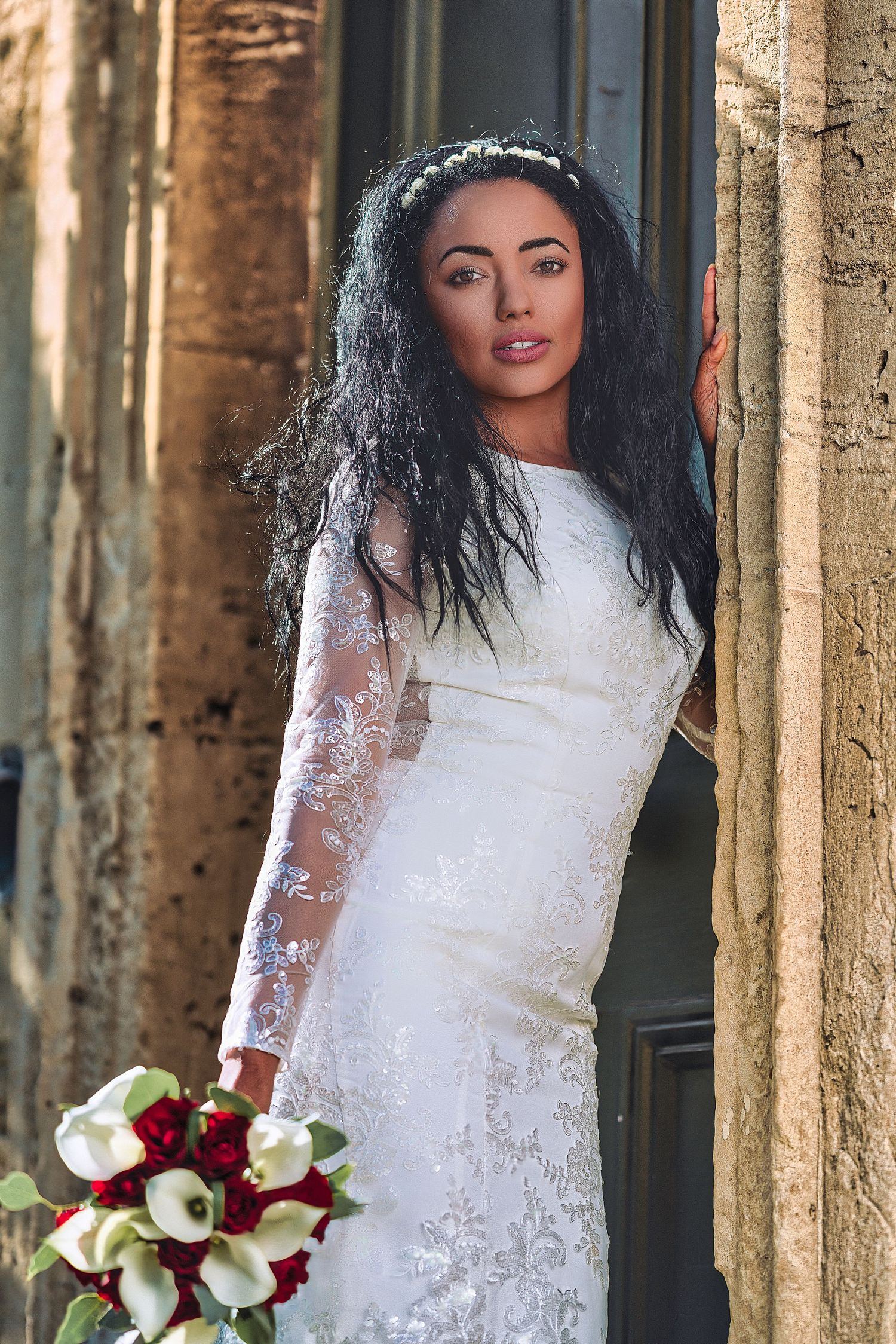 Bride in lace gown holds red and white bouquet against stone columns.