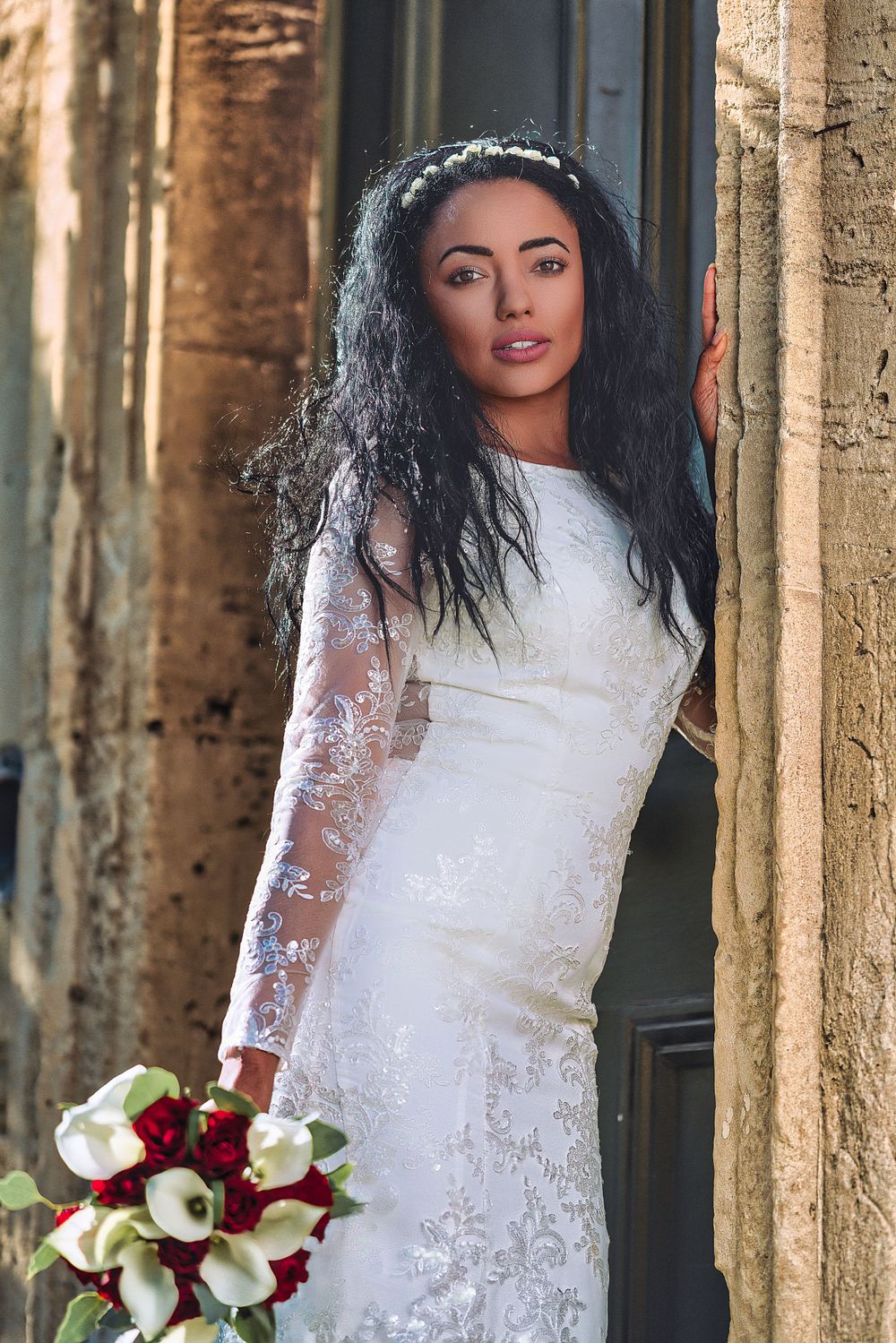 Bride in lace gown holds red and white bouquet against stone columns.