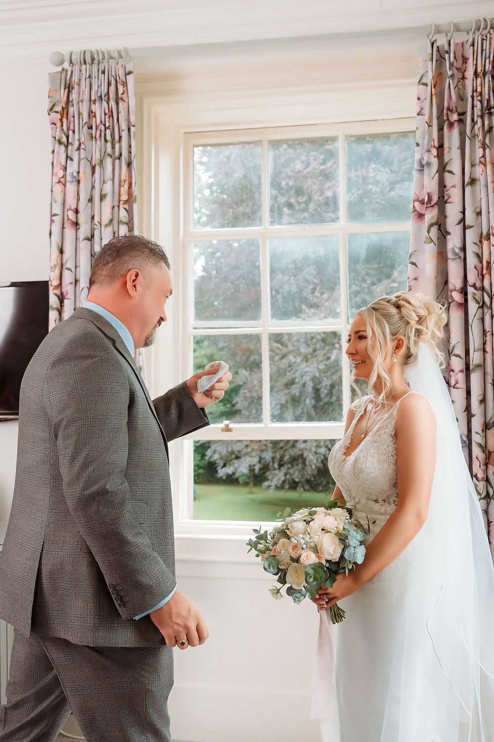 A couple shares a tender moment by a bright window with floral curtains during a wedding ceremony.