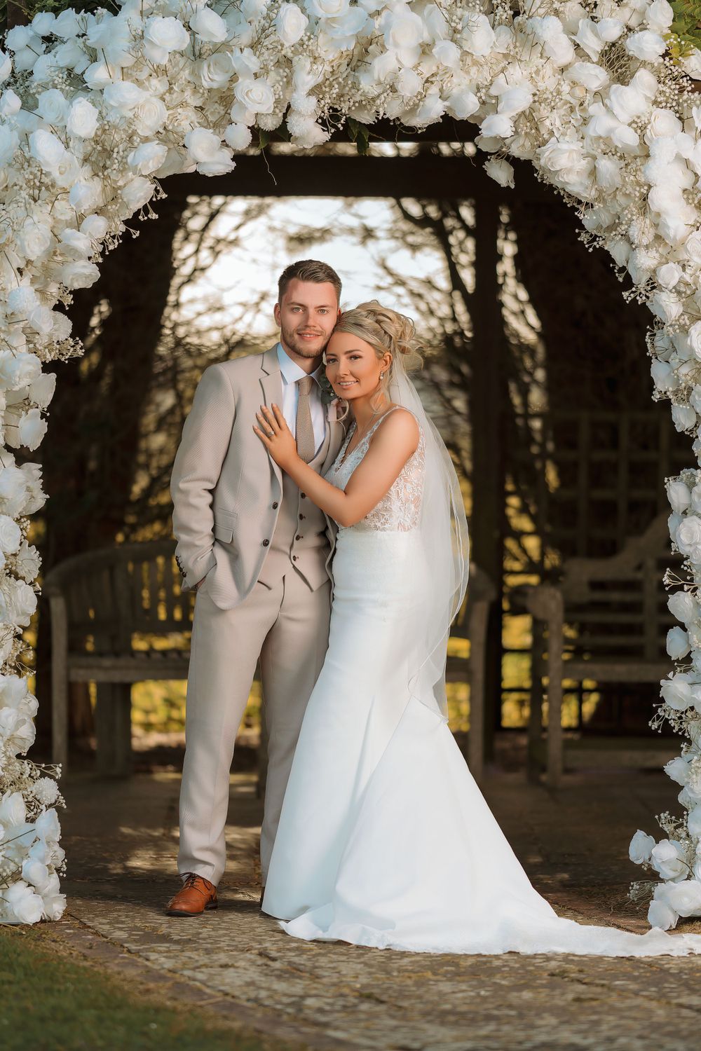 Romantic wedding photos captured under a white floral arch in an outdoor garden setting during golden hour.