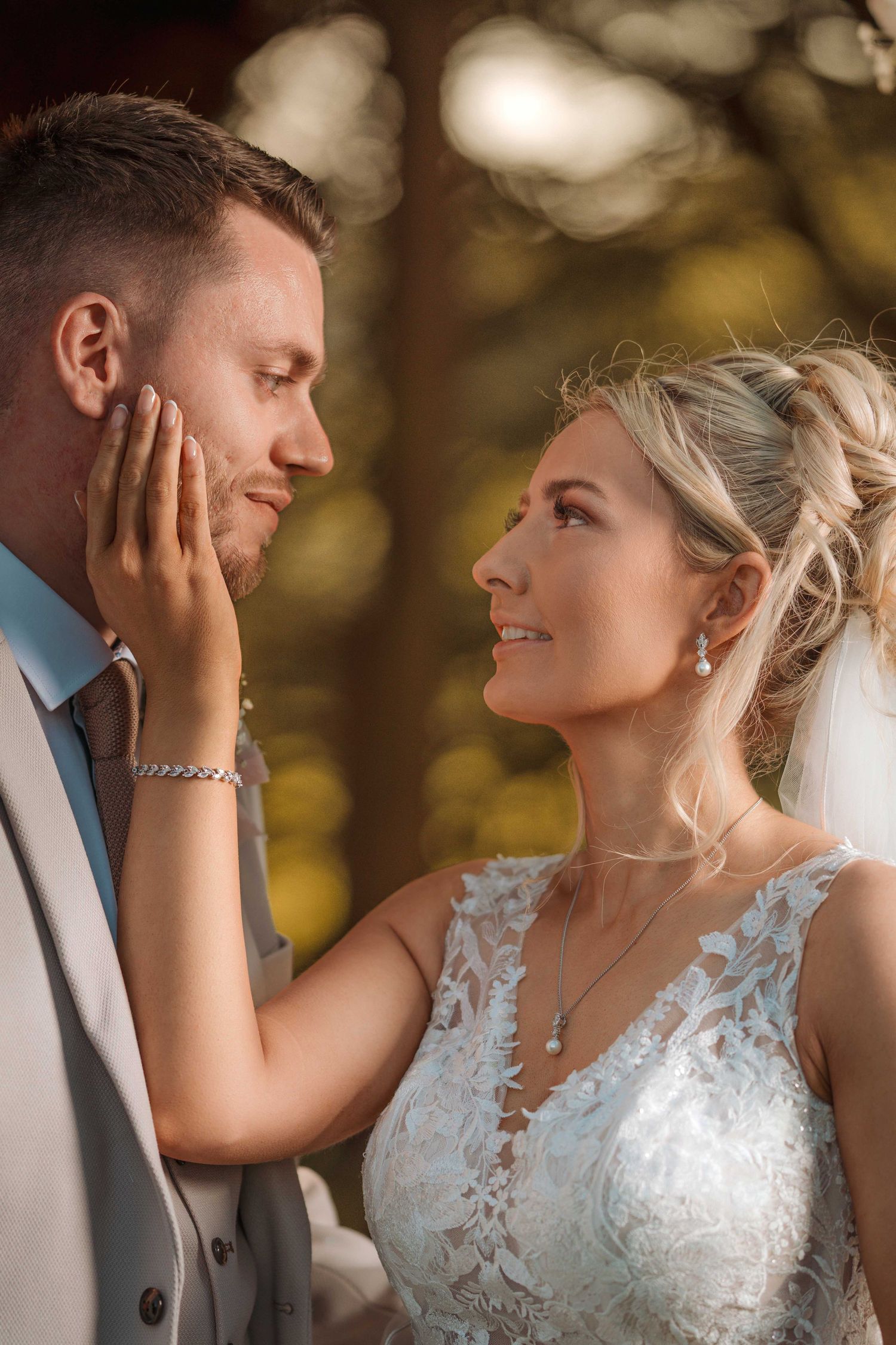 Bride in lace gown tenderly cups groom's face as they share an intimate moment outdoors in warm golden light.