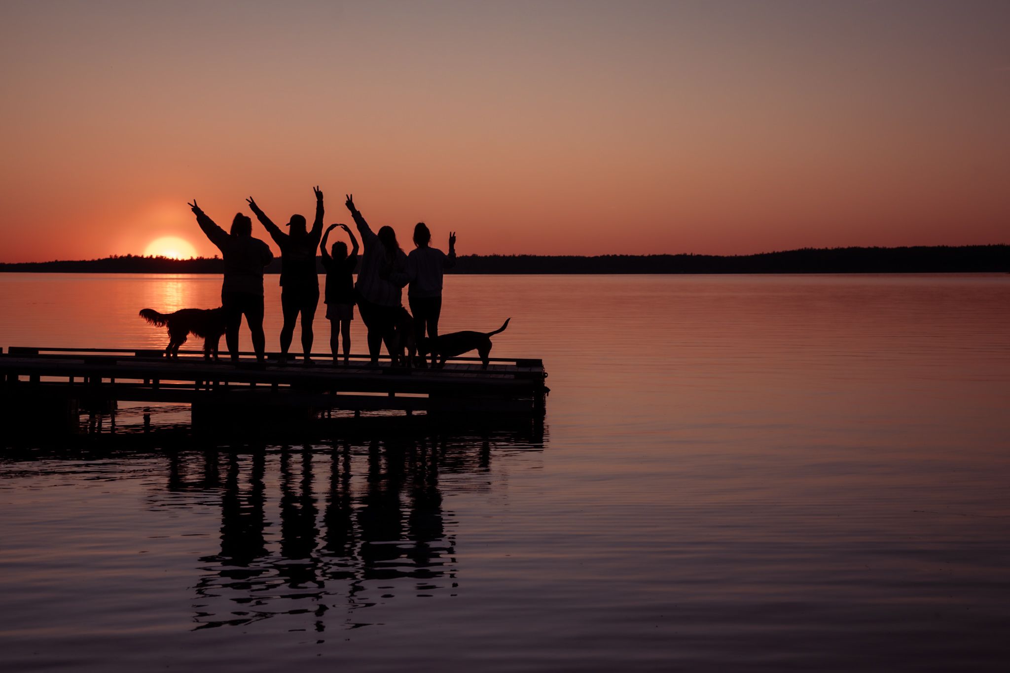 Silhouettes of people celebrating on a dock at sunset with reflections in the calm lake water.