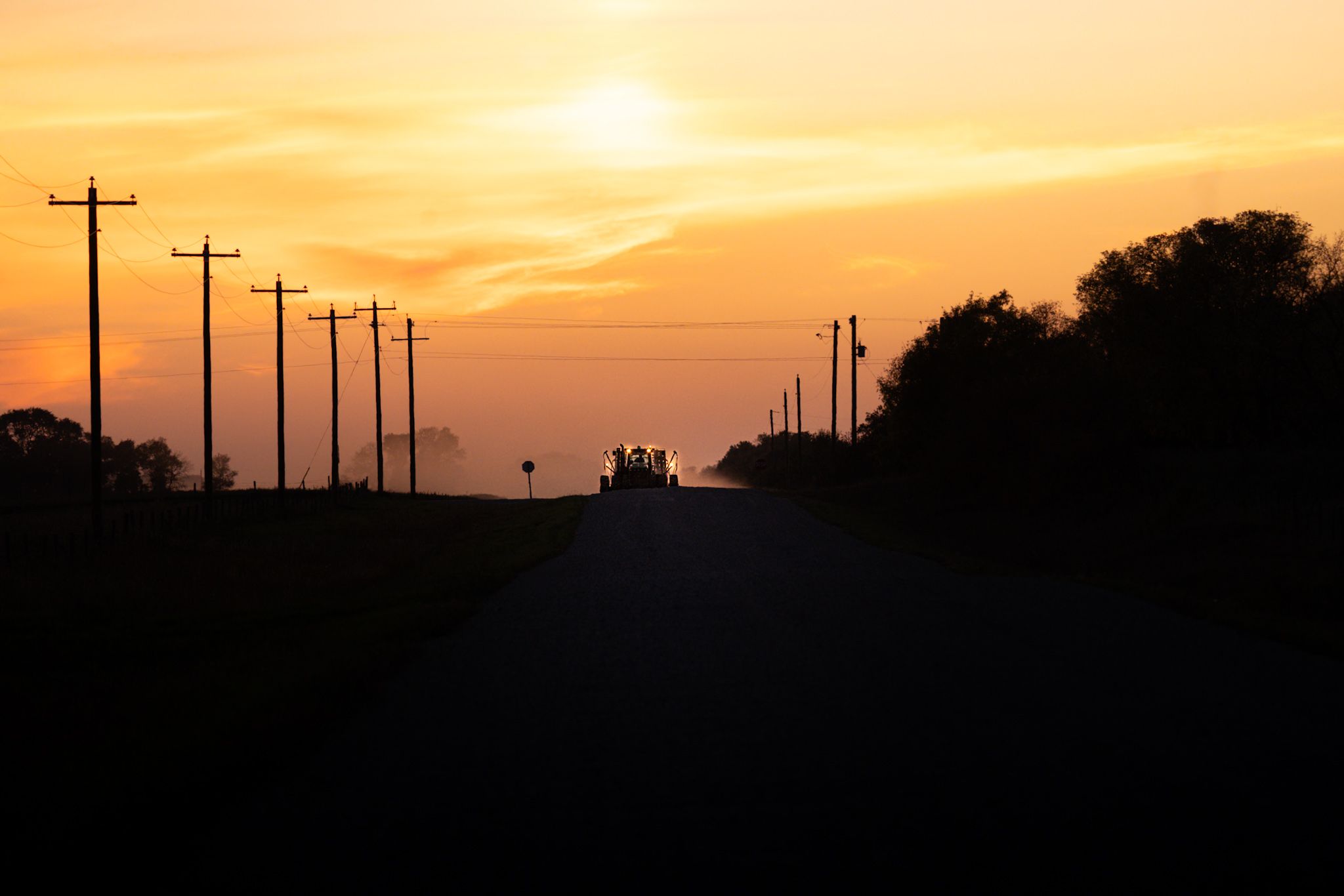 Silhouette of utility poles line a rural road against a dramatic orange sunset sky.