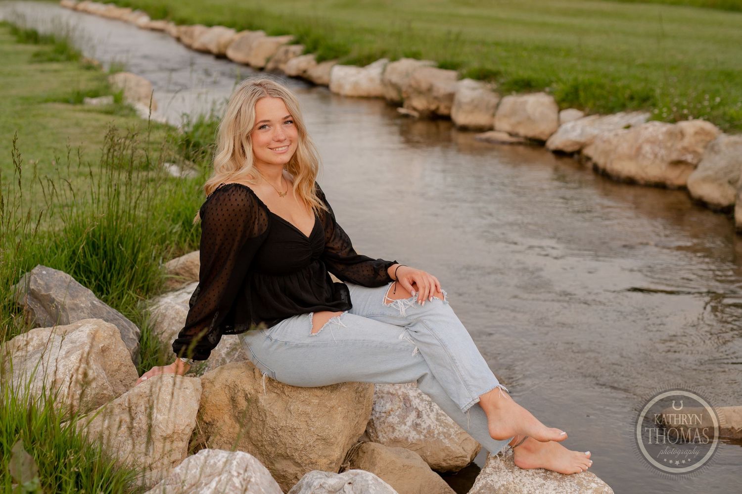 Portrait sitting on rocks along flowing stream wearing black top and jeans.