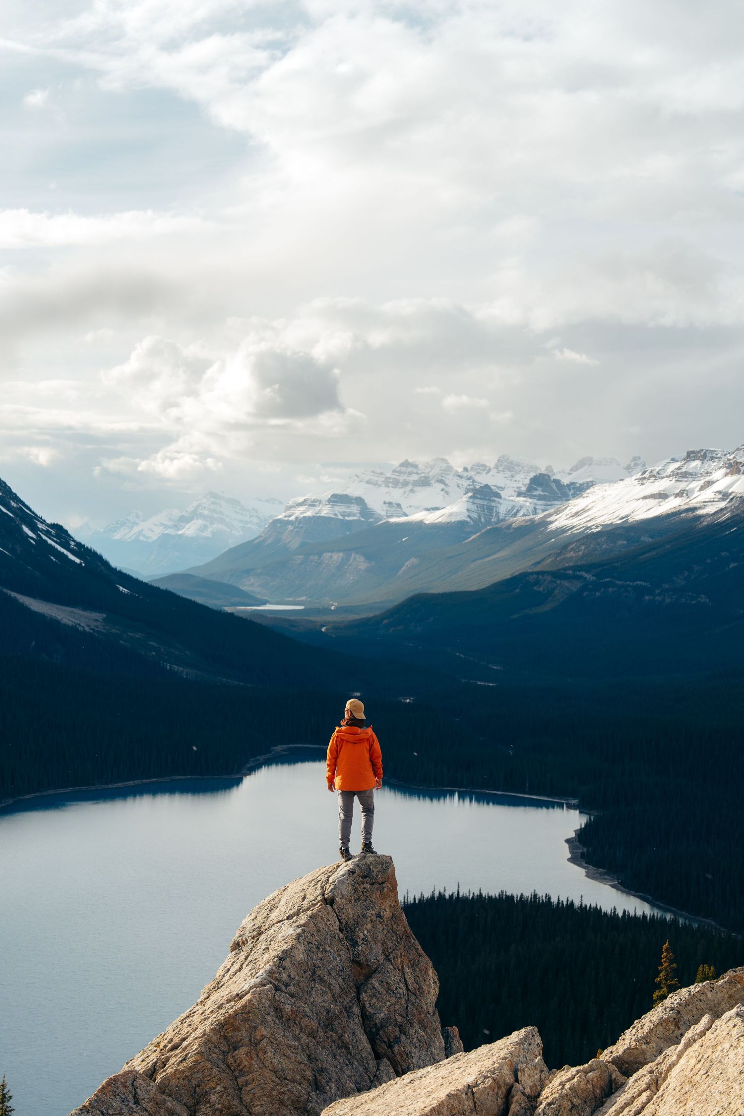 A hiker in an orange jacket stands on a rocky cliff overlooking a serene mountain lake surrounded by snowy peaks.