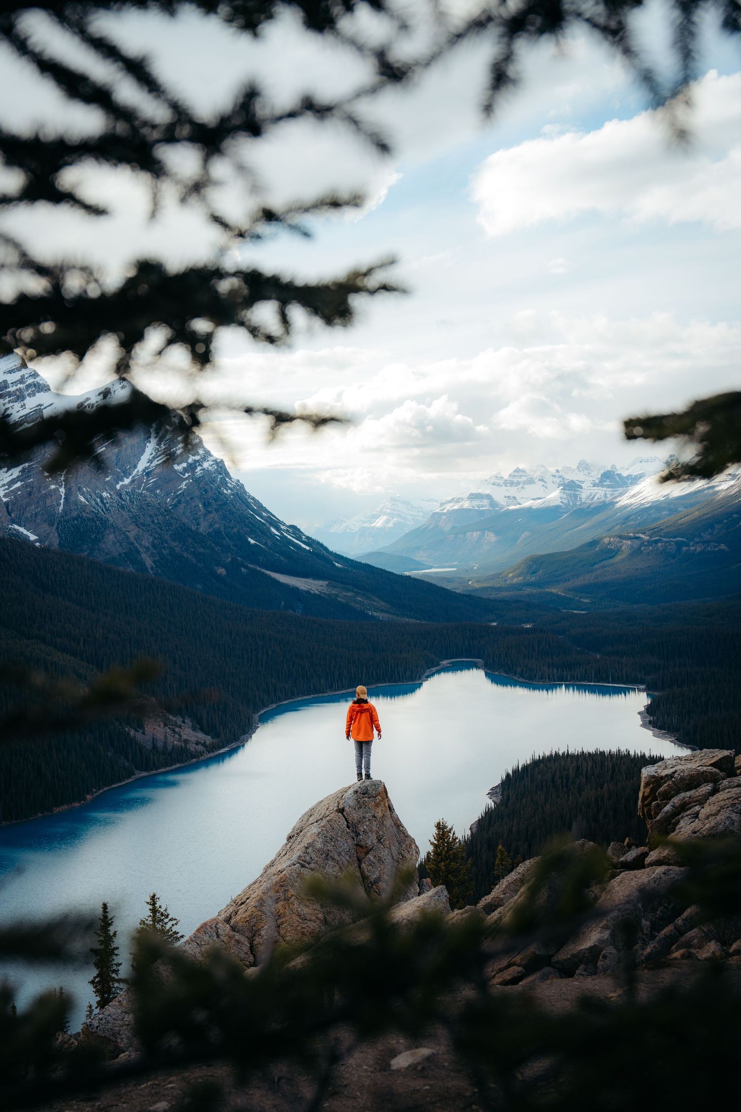 Adventurer in orange jacket stands on mountain overlook above serene lake with snowy peaks and forests in Canadian Rockies.