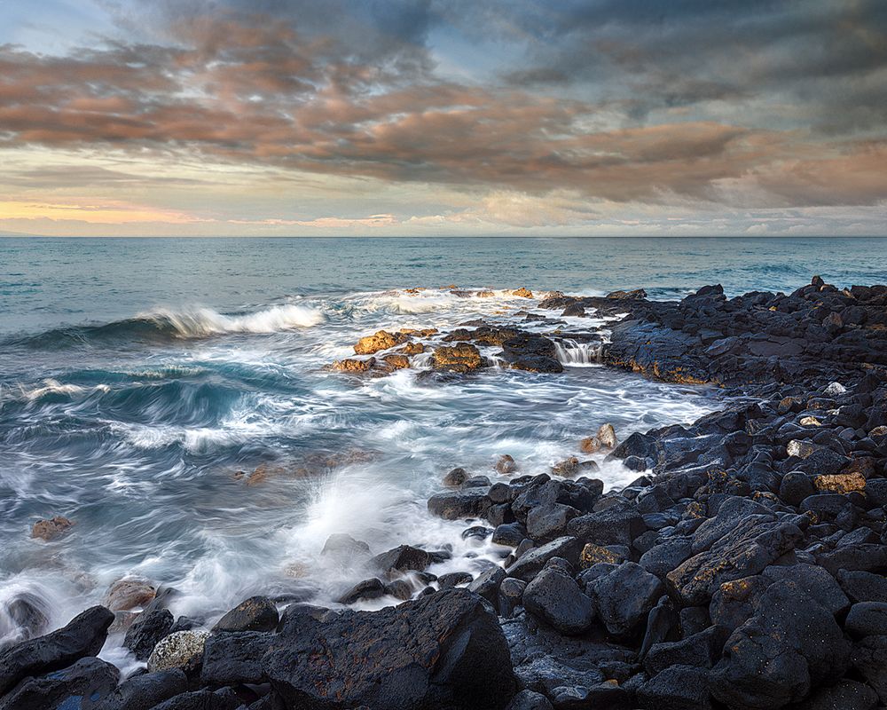 Rocky coastline at sunset with waves crashing against dark stones under colorful clouds.