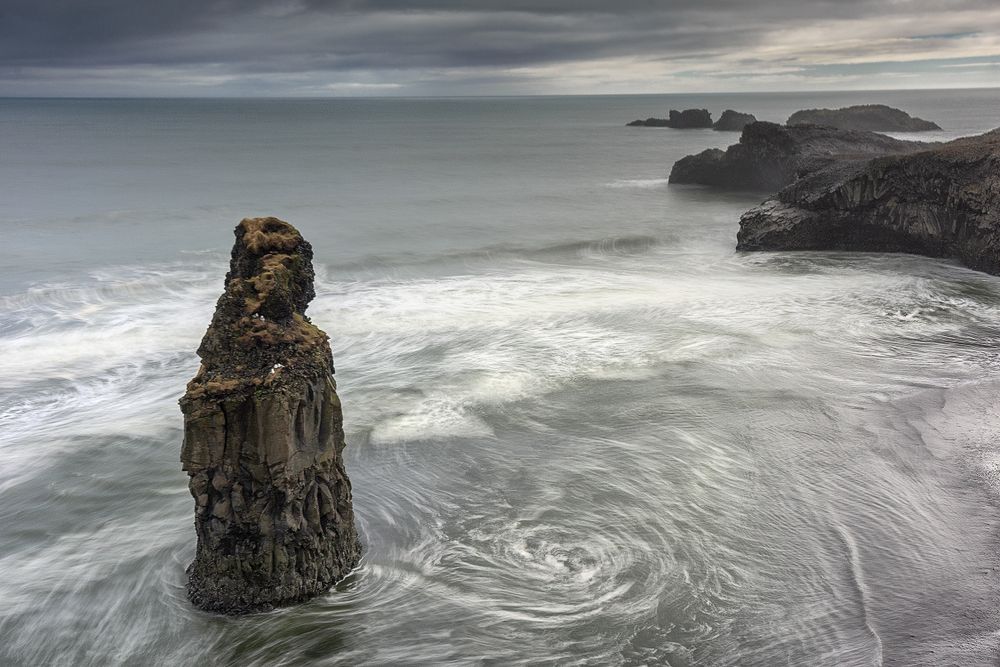 Lone sea stack standing in swirling ocean waters against a moody grey sky.
