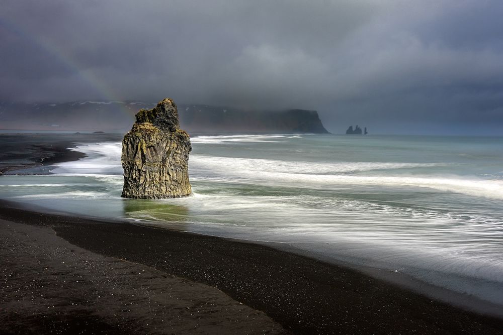 Isolated sea stack on black sand beach with misty cliffs in the distance.