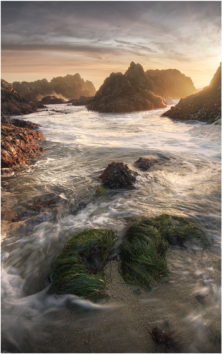 Turbulent seawater flows through rocky coastal formations during dramatic golden sunset.